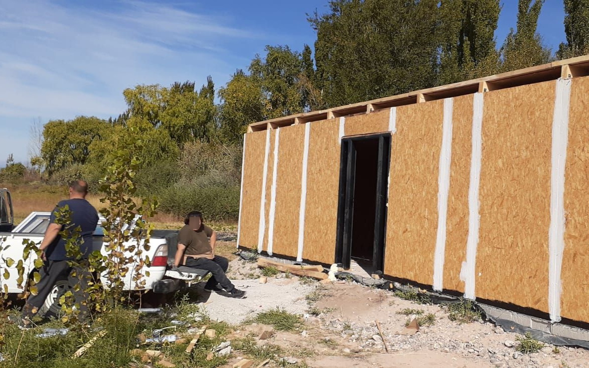 Exterior de un edificio en construcción. Dos personas cerca de una camioneta blanca. Paredes de madera con marco de puerta.