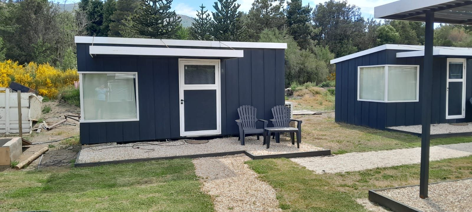Dos pequeñas cabañas de color azul oscuro con detalles blancos, dos sillas y caminos de grava en un entorno verde.