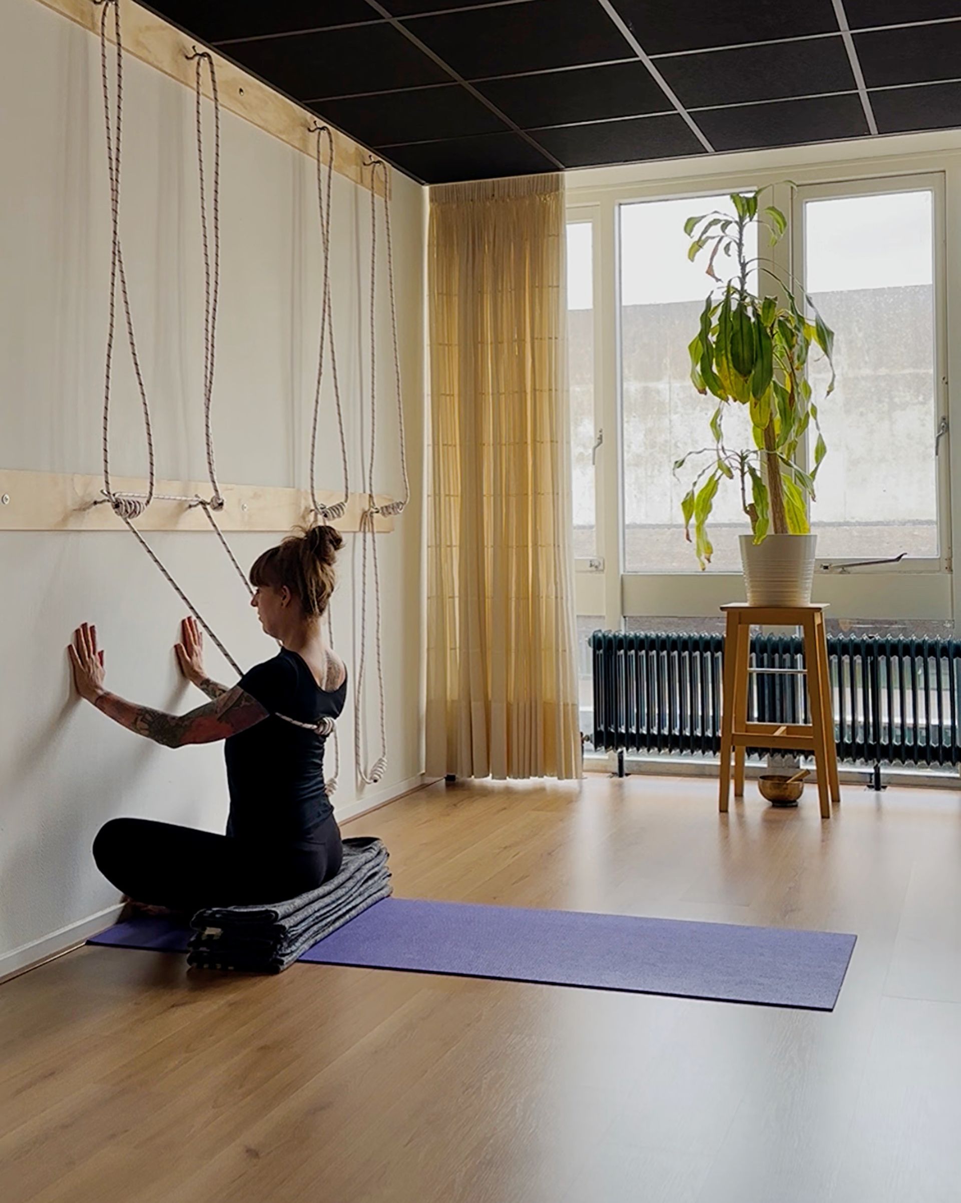 Baddha konasana, zittend op yoga dekens bij de muur, met een touw om de romp dat steunt de tilling  ervan.
