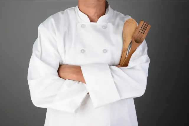 Chef in white uniform with arms crossed, holding wooden utensils.