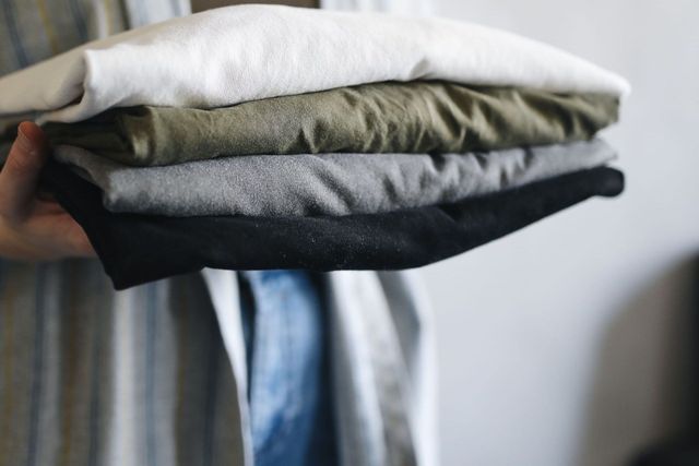 Person holding a stack of folded t-shirts: white, olive, gray, and black.