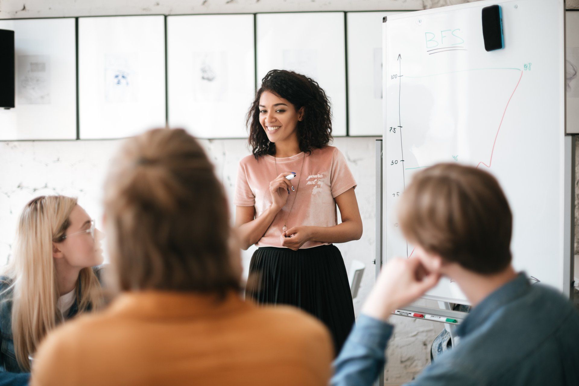 Portrait of cheerful African American lady with dark curly hair standing near board and happily speaking with colleagues in office. Young beautiful business woman giving presentation to coworkers