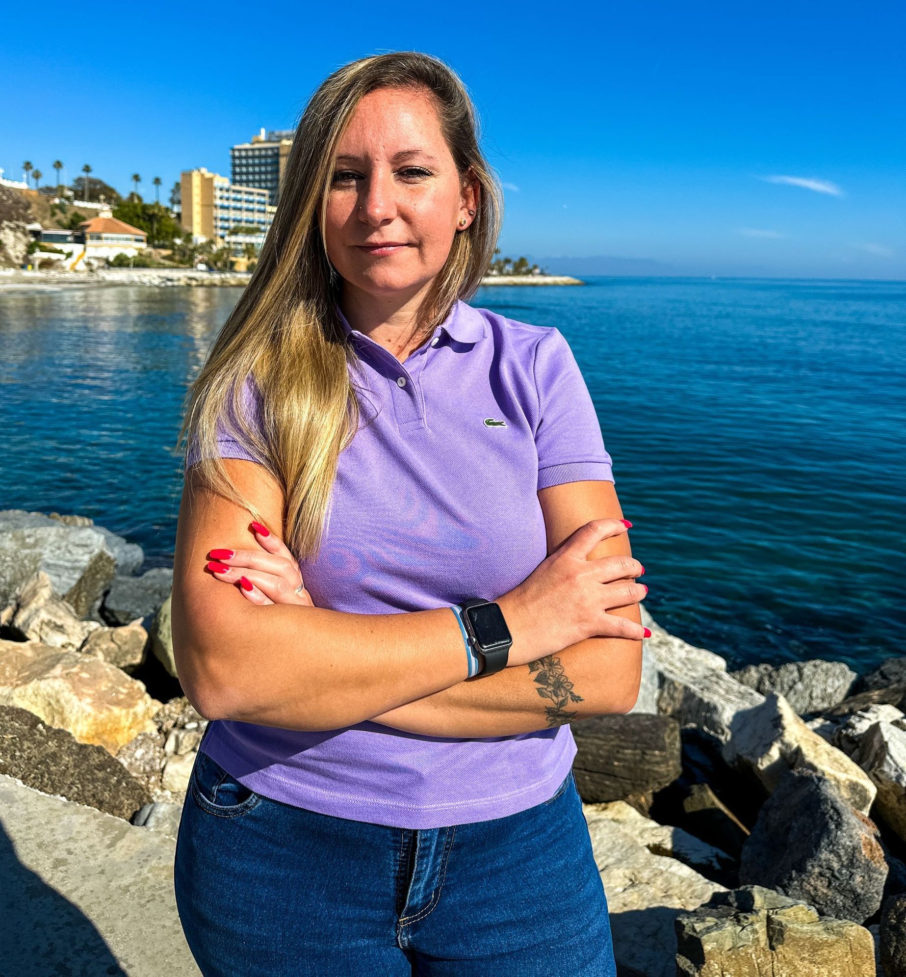 Una mujer con una camisa morada está parada junto al océano con los brazos cruzados.