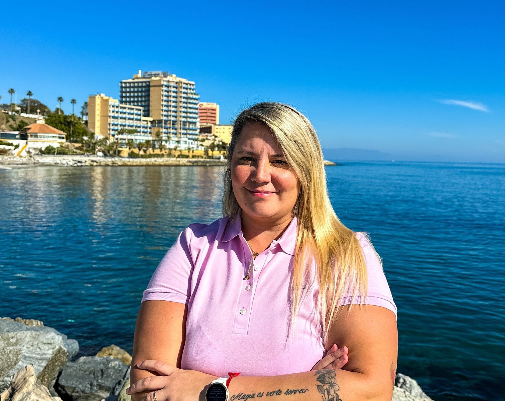 Una mujer con una camisa rosa está parada sobre una roca cerca del océano.