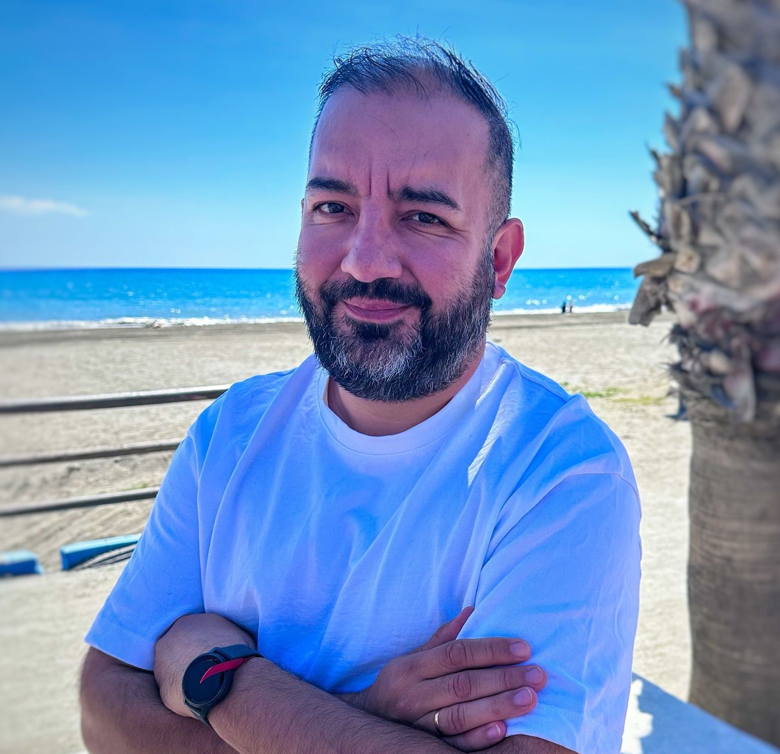 Un hombre con barba está de pie en una playa con los brazos cruzados.