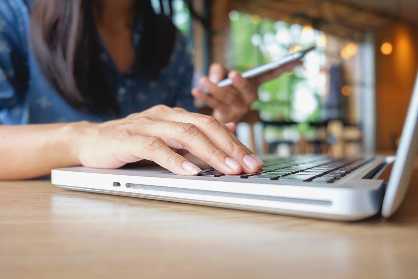 Una mujer está escribiendo en una computadora portátil mientras sostiene un teléfono celular.
