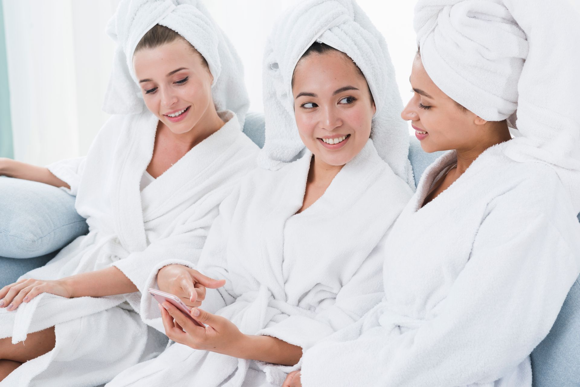 Three women in bathrobes and towels on their heads are sitting on a couch looking at a cell phone.