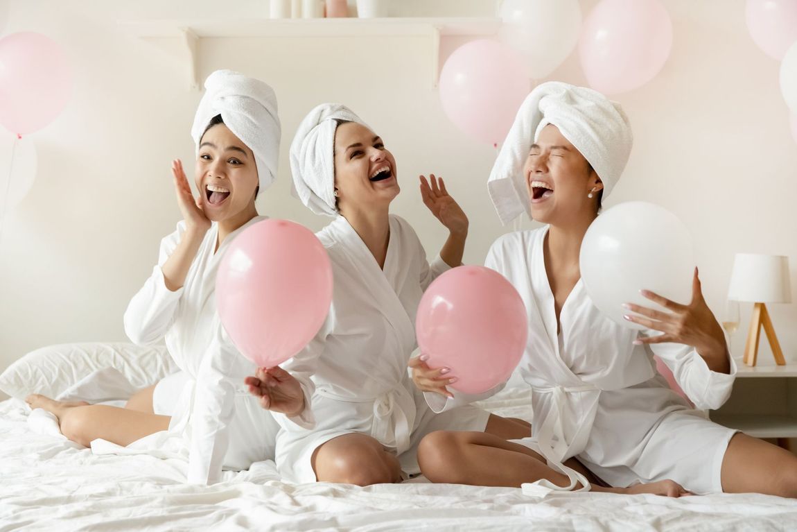 Three women with towels wrapped around their heads are sitting on a bed holding pink balloons.