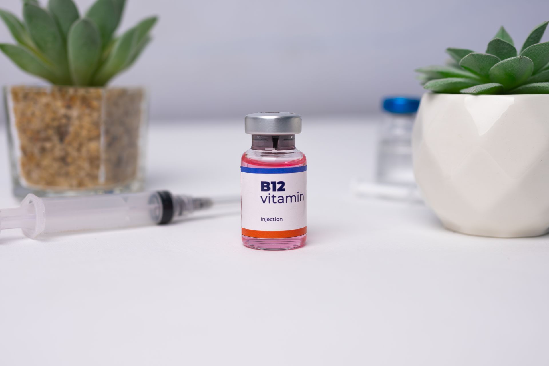 Vial of B12 vitamin with a syringe, succulents in pots, on a white surface.
