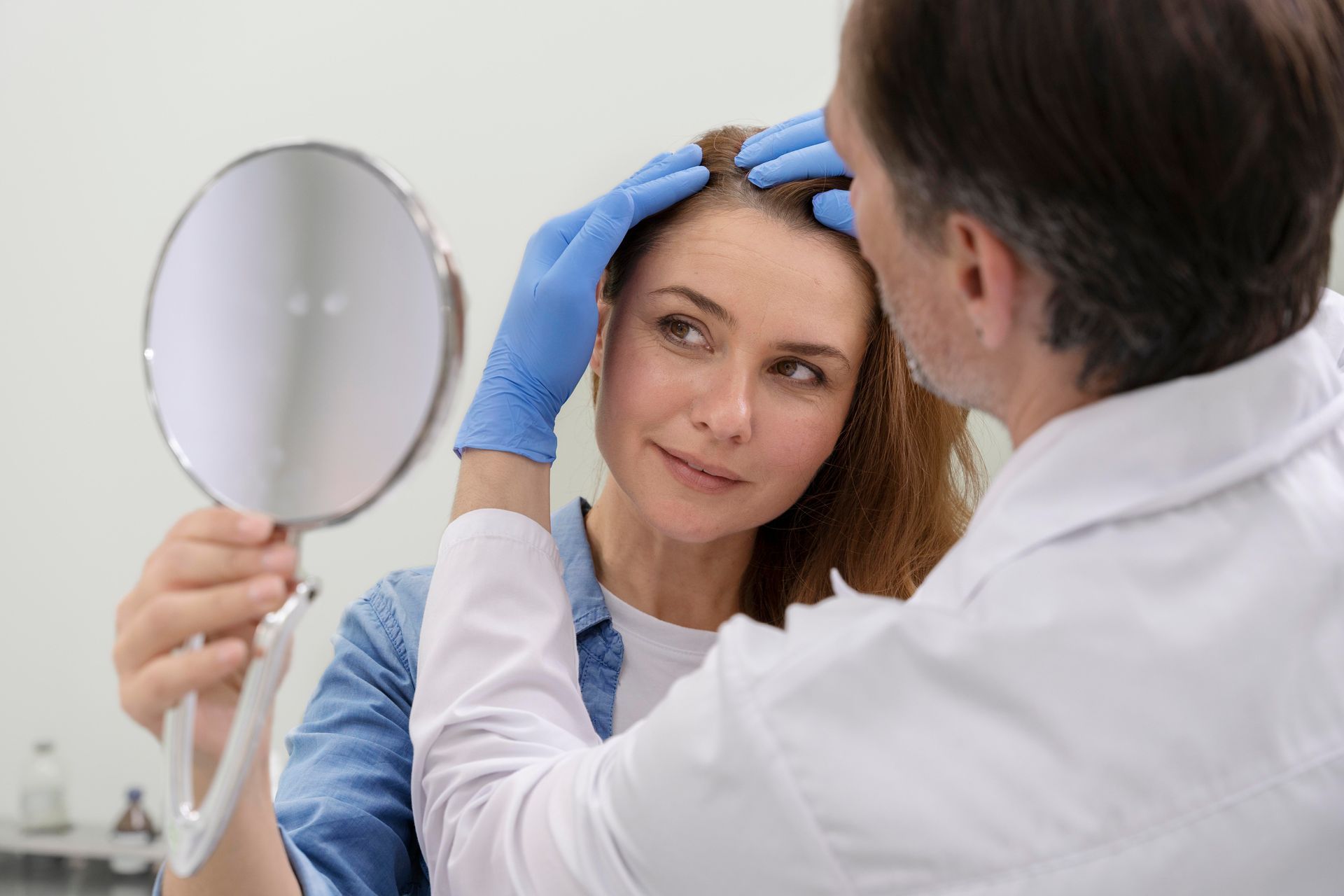 A doctor in blue gloves examines a woman's scalp with a hand mirror in a clinic.