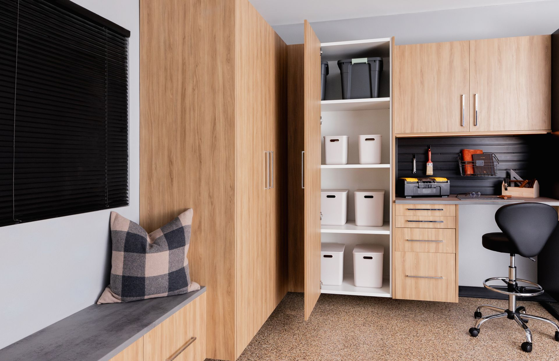 A garage Cabinet featuring light wood cabinetry, white storage bins, a desk with a black rolling chair, and a window seat.