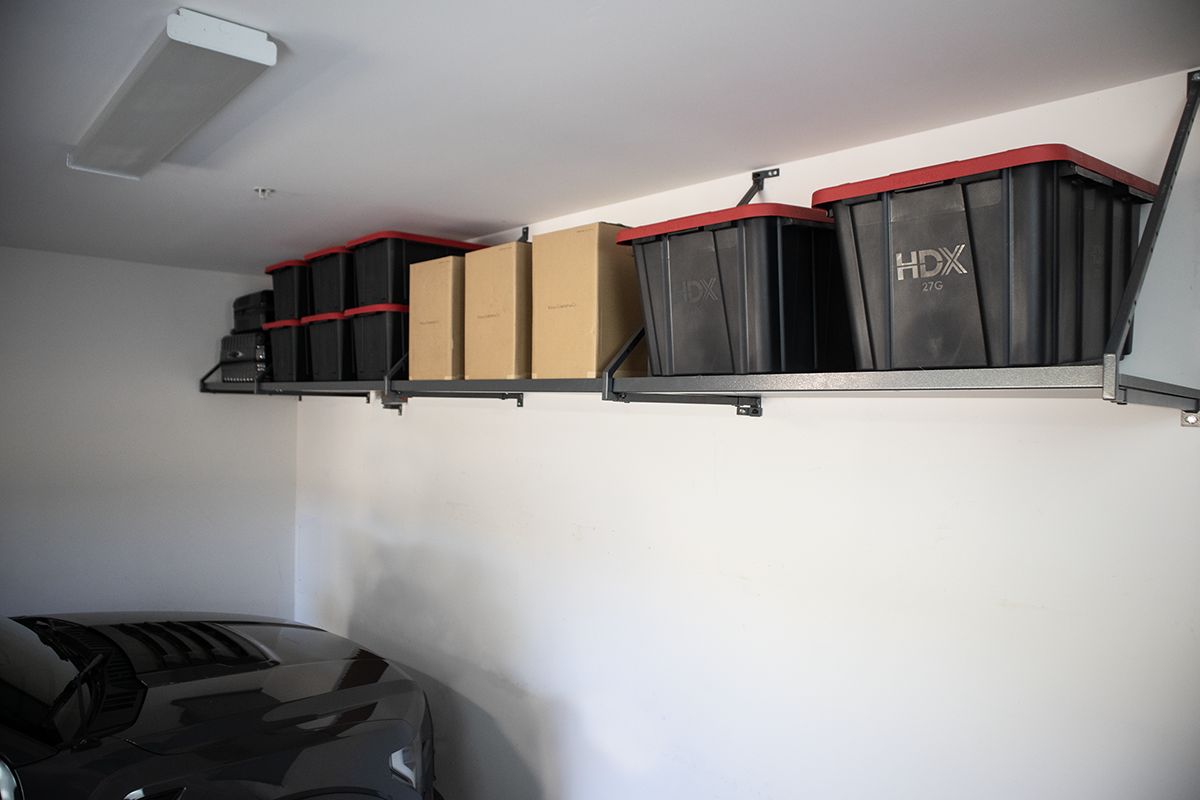 Overhead storage rack mounted in a garage holding black plastic bins and cardboard boxes above a parked car.