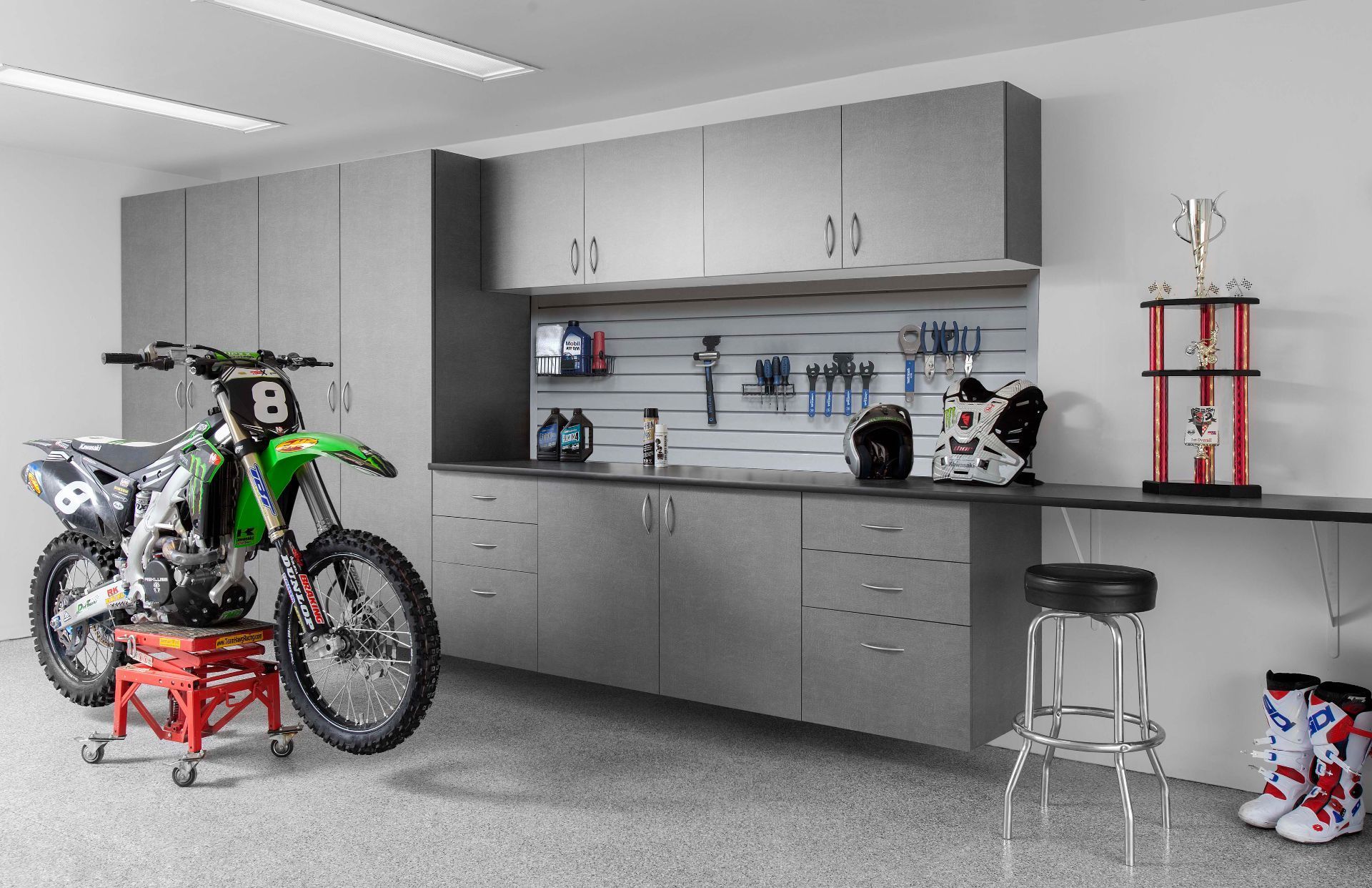A green motocross bike on a stand in a garage with grey cabinetry, a workbench, and a trophy on display.