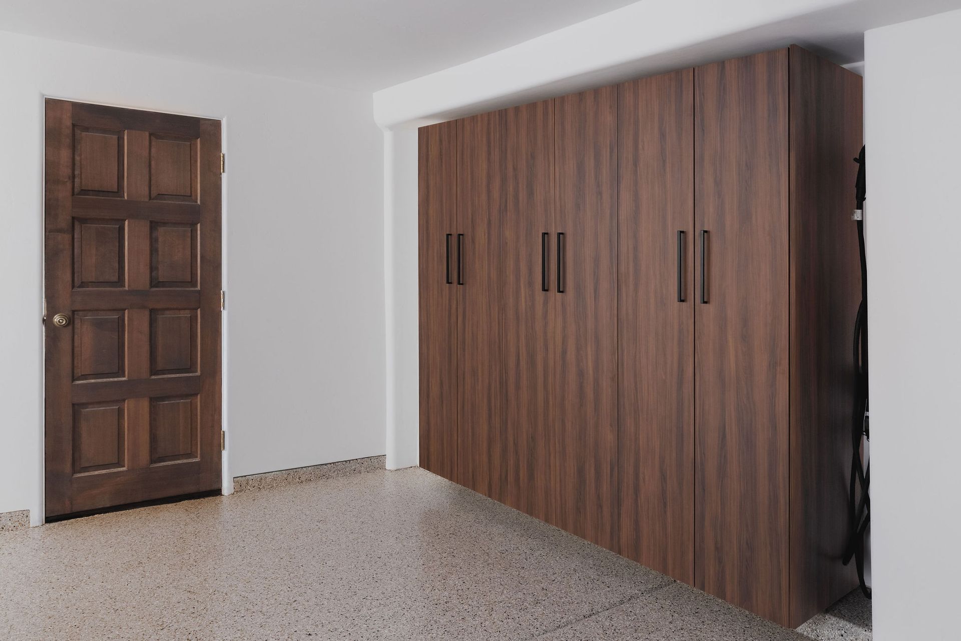 A brown wood-paneled storage cabinet sits against a white wall near a matching wood door in a room with speckled flooring.