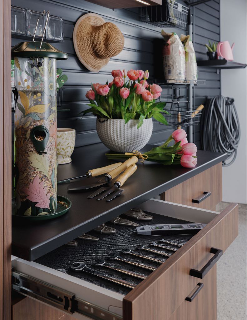 A dark wood cabinet drawer open to reveal garden tools, with a potting station above holding flowers and a bird feeder.