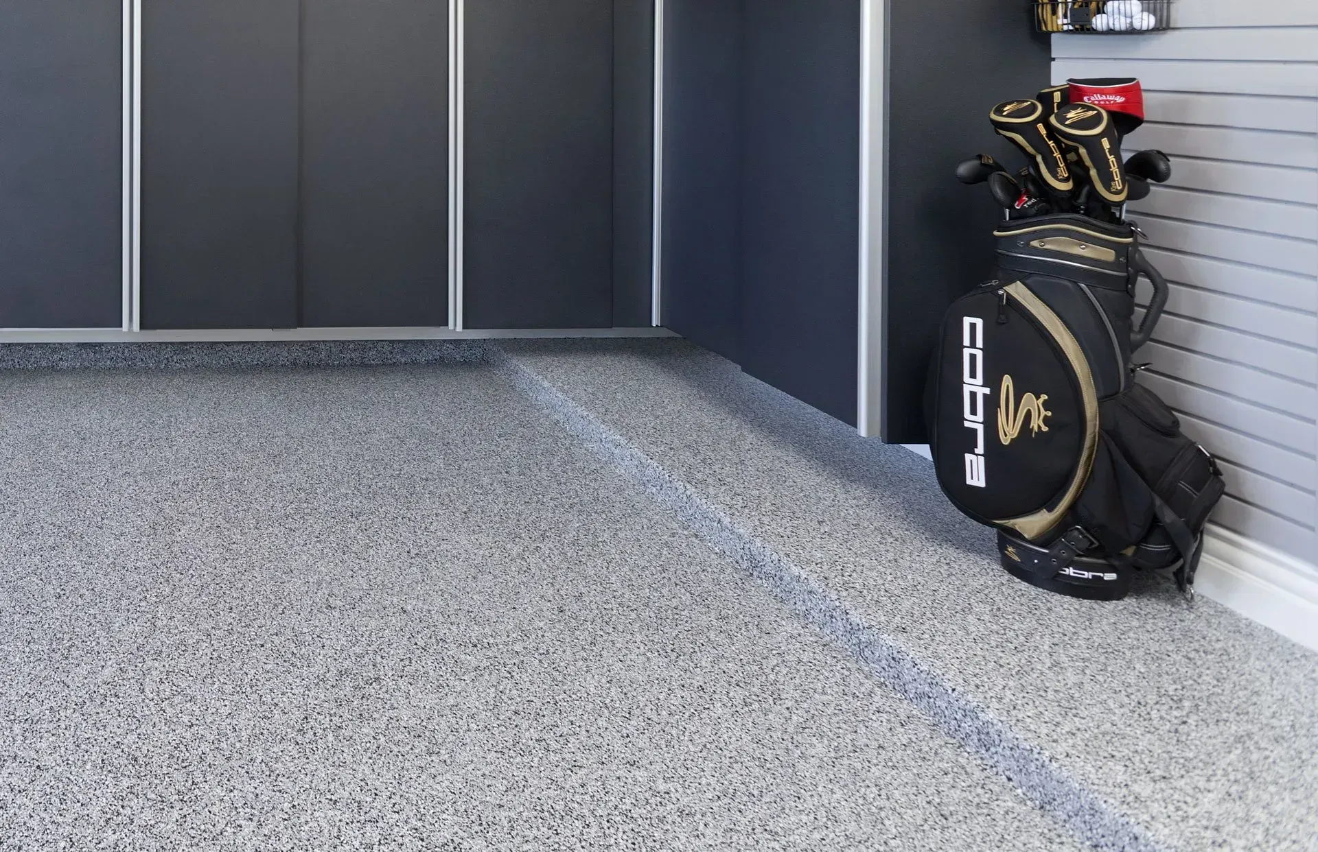 A Cobra golf bag stands in a garage with grey speckled epoxy flooring and dark cabinets.