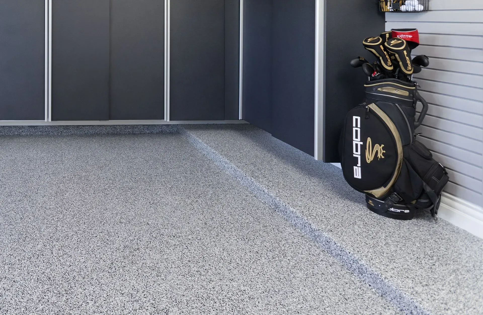 A black Cobra golf bag stands in the corner of a garage with dark cabinets and a grey speckled epoxy floor.