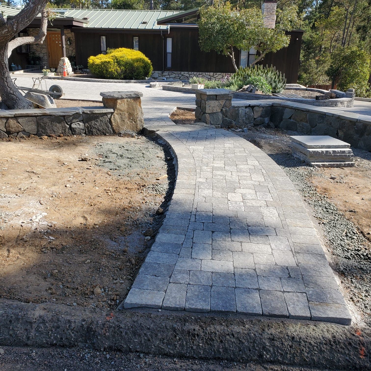 A stone walkway leads toward a dark-sided house with a green roof, framed by low stone retaining walls and dirt landscaping.