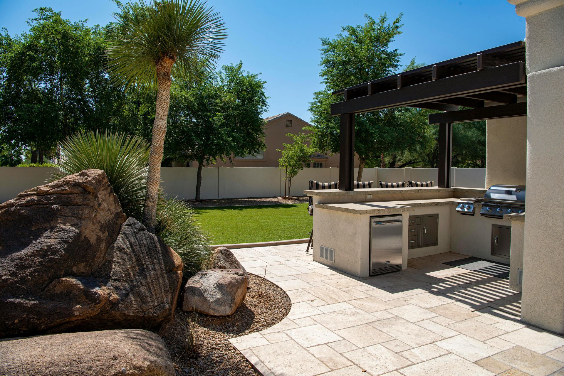 Backyard patio with outdoor kitchen, grill, and pergola, with green lawn and large rocks.