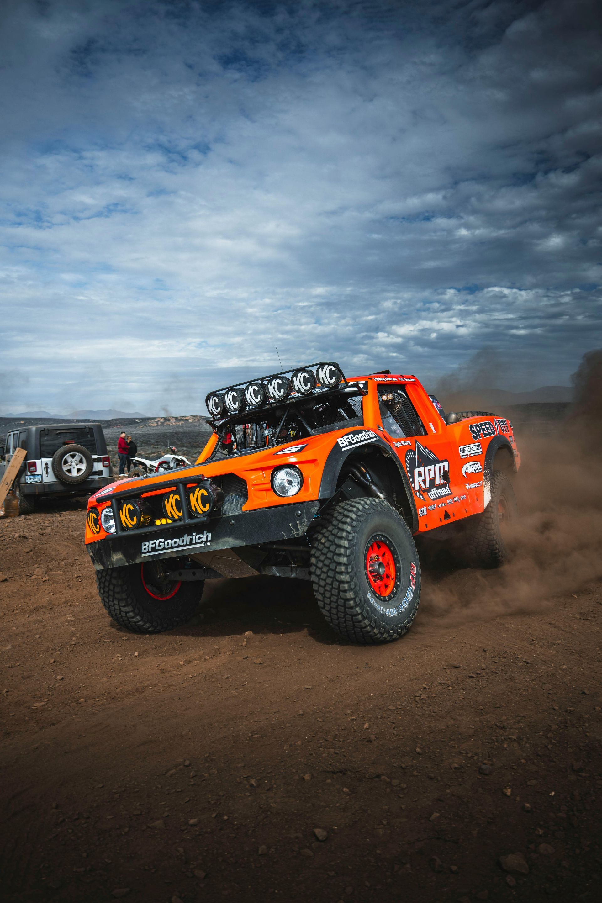 Orange off-road truck speeding on a dirt track, kicking up dust. A second vehicle and cloudy sky are in the background.