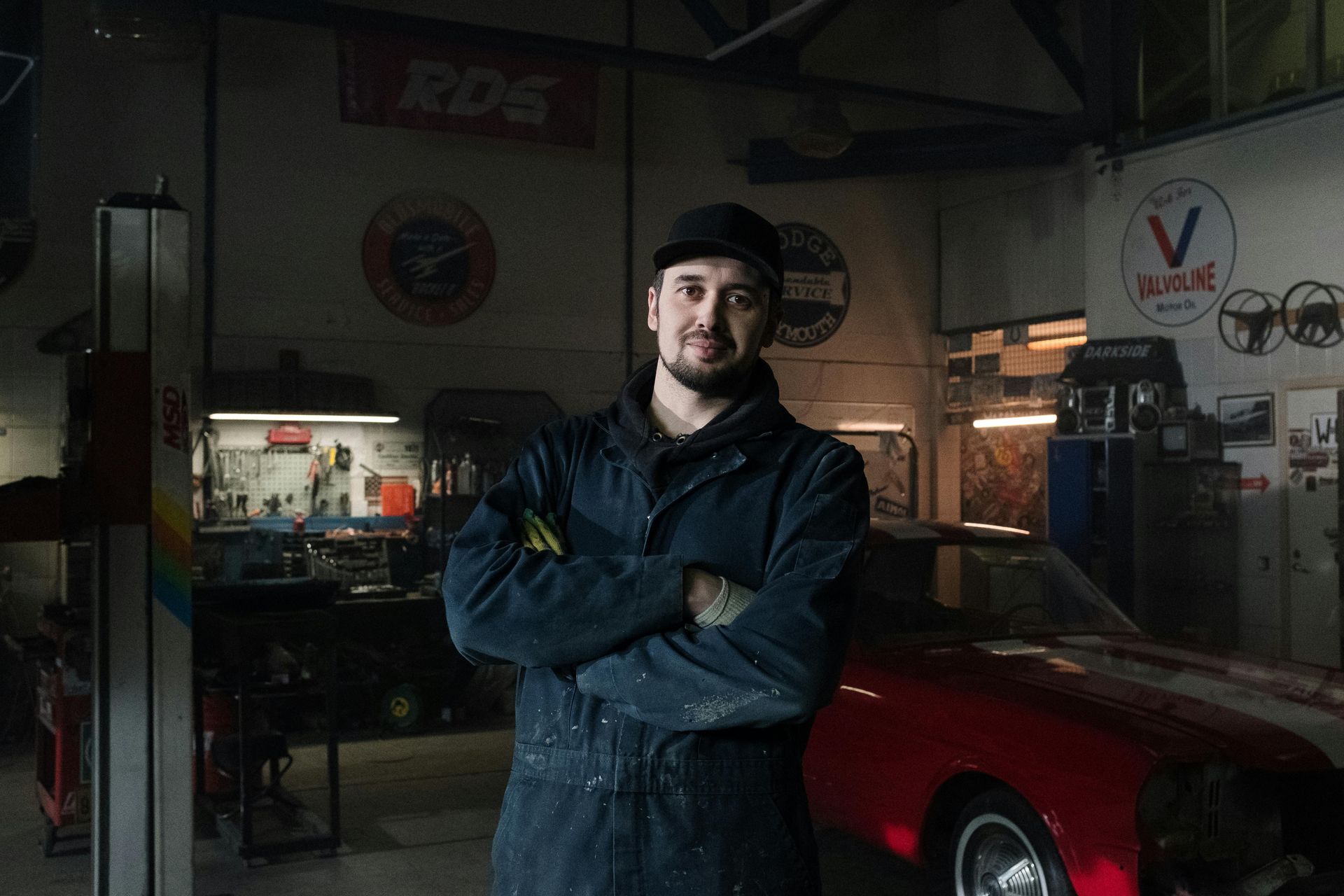 Mechanic in a blue jumpsuit stands in a garage with arms crossed, red sports car to his right.