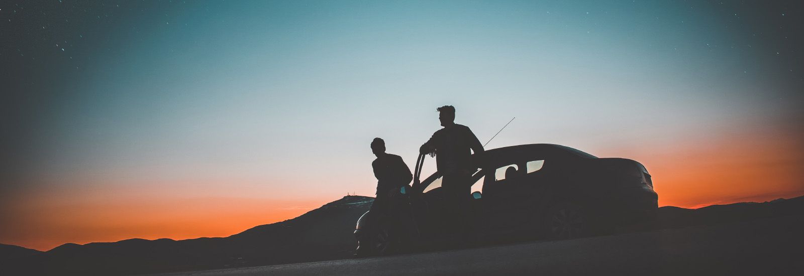 Silhouetted figures beside a vehicle against an orange and blue sunset sky.