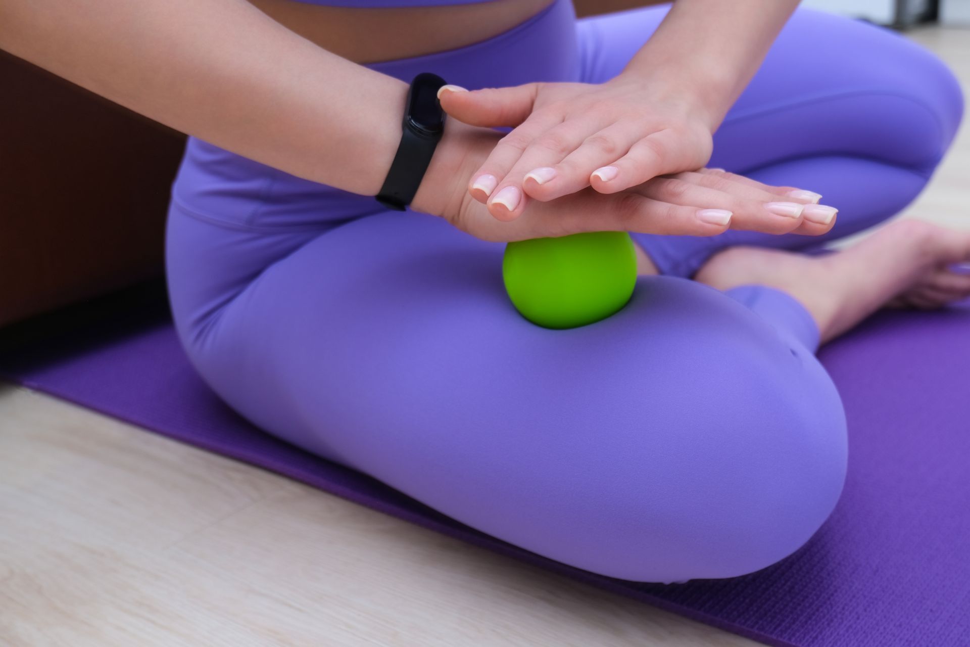 A woman is sitting on a yoga mat with a green ball on her leg.