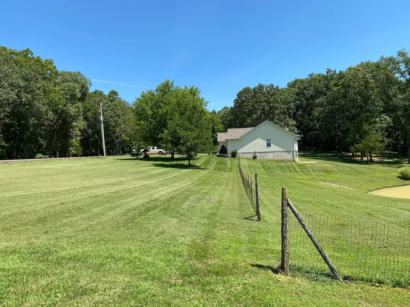 A house sits amid a large, mowed grassy area, with a wooden fence in the foreground and trees on the horizon under a blue sky.