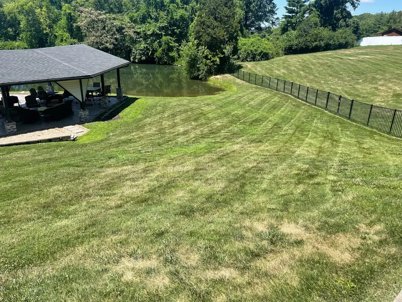 Green lawn slopes to a pond, with a covered patio on the left and a black fence on the right.