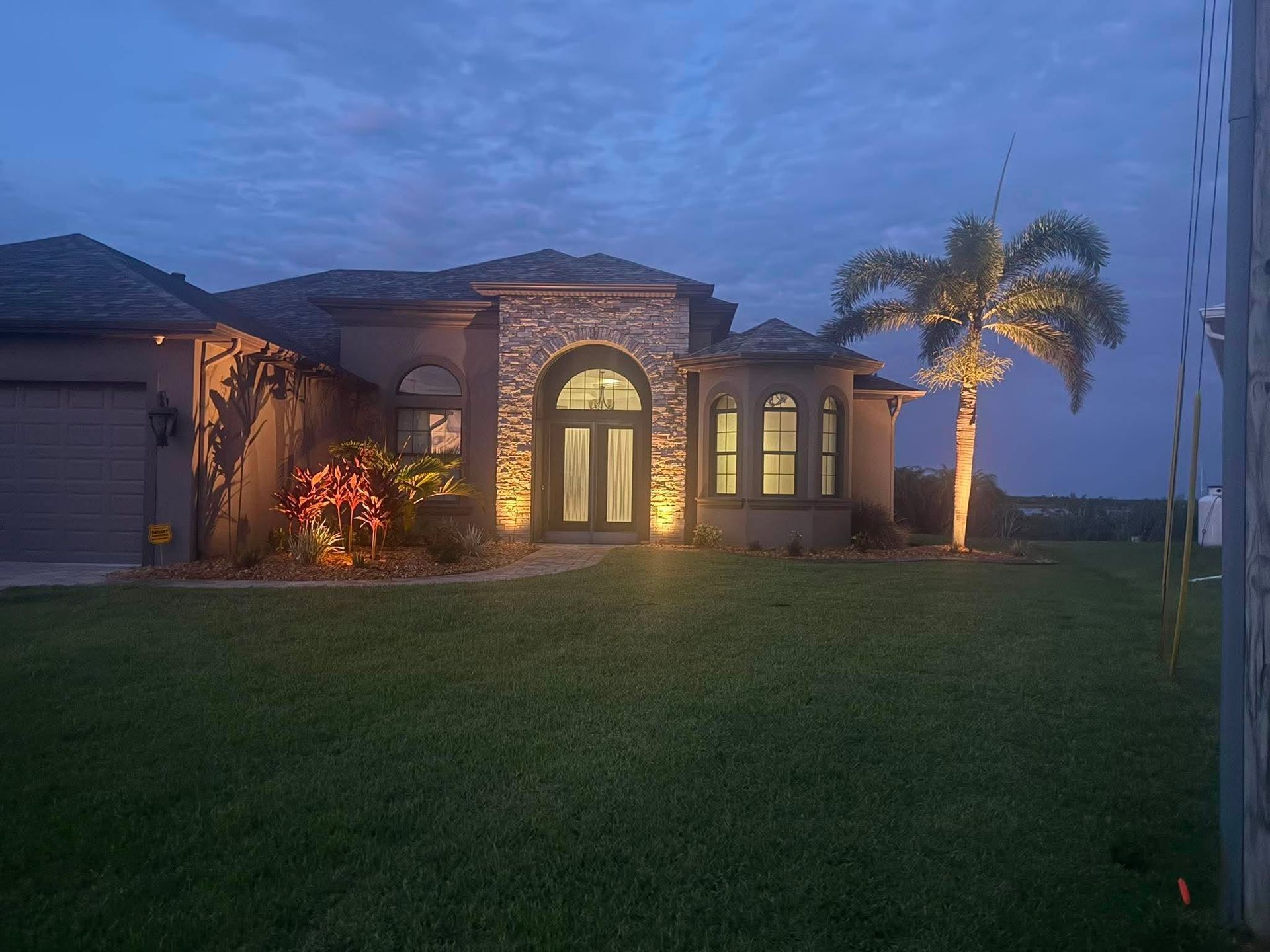 Twilight view of a single-story house with lit entryway, stone facade, palm tree, and large lawn.