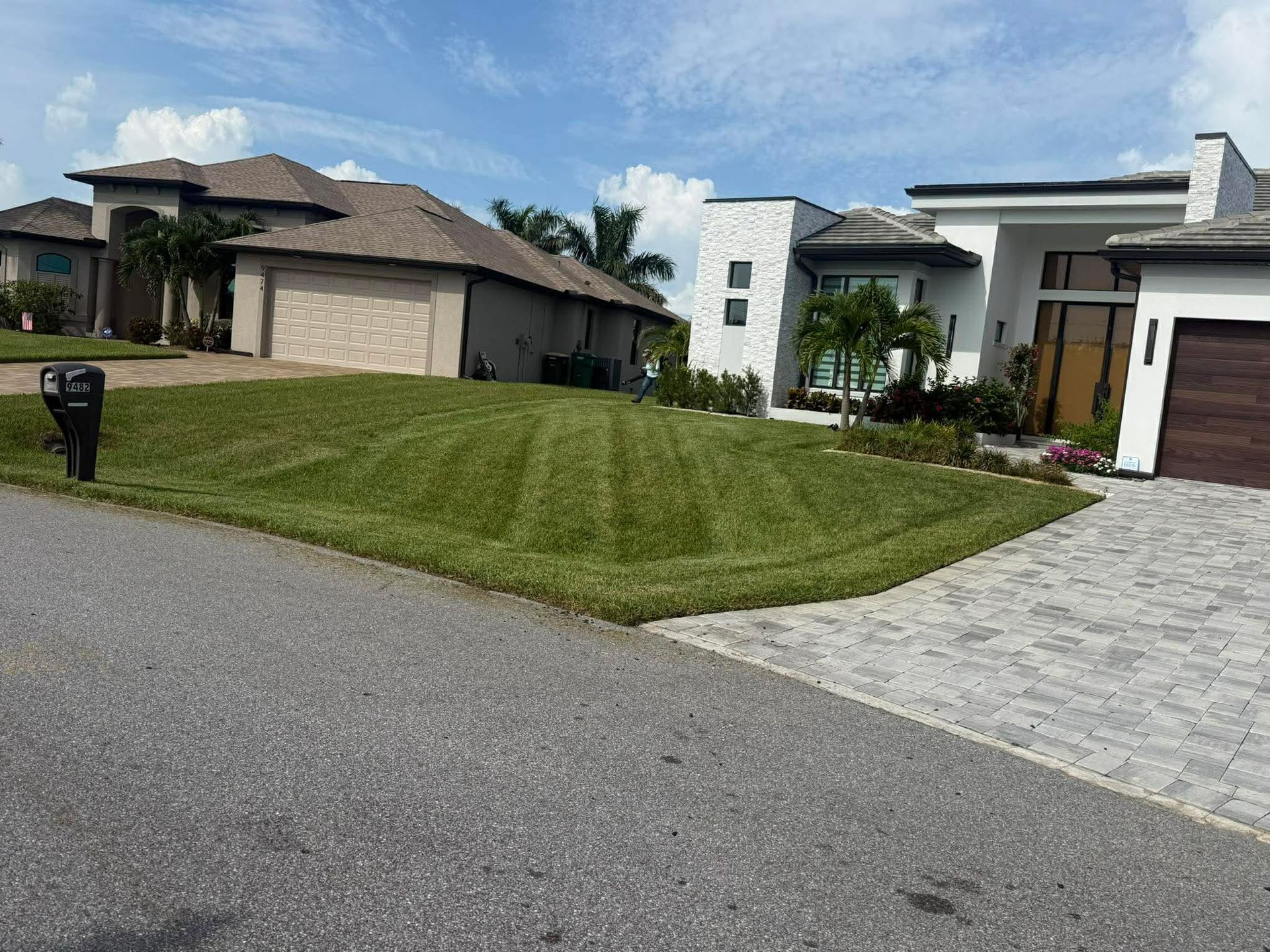 Suburban street with modern houses, a grassy lawn, and a paved driveway under a blue sky