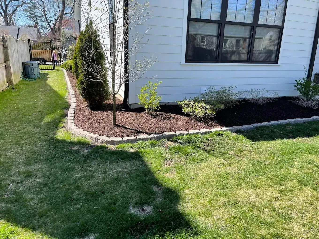 A house corner with a mulch-covered garden bed, bordered by brick, next to a grassy lawn.