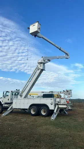 Bucket truck with extended arm reaching up to power lines, against a cloudy sky.