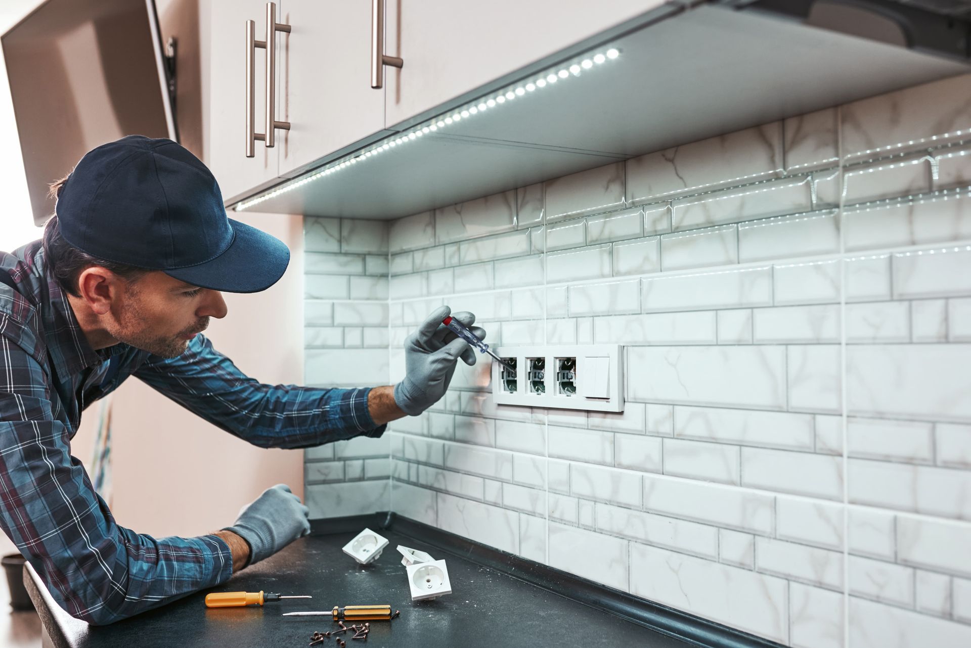 A person installing an electrical outlet in a kitchen. They are wearing gloves and a cap.