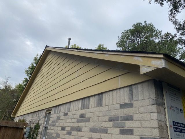 Yellow-sided house with a dark roof and brick foundation under a cloudy sky.