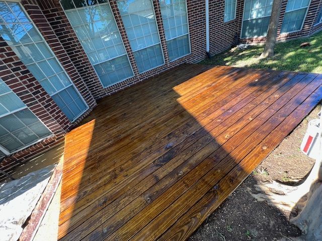 Wooden deck attached to a brick house. The deck has a fresh stain, with bright sunlight reflecting on it.