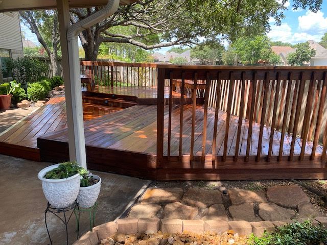 Wooden deck with dark stain, surrounded by a yard with plants, trees, and a light-colored patio.