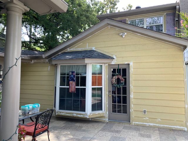 Yellow house with bay window, door, and patio. American flag hanging in the window, wreath on the door.