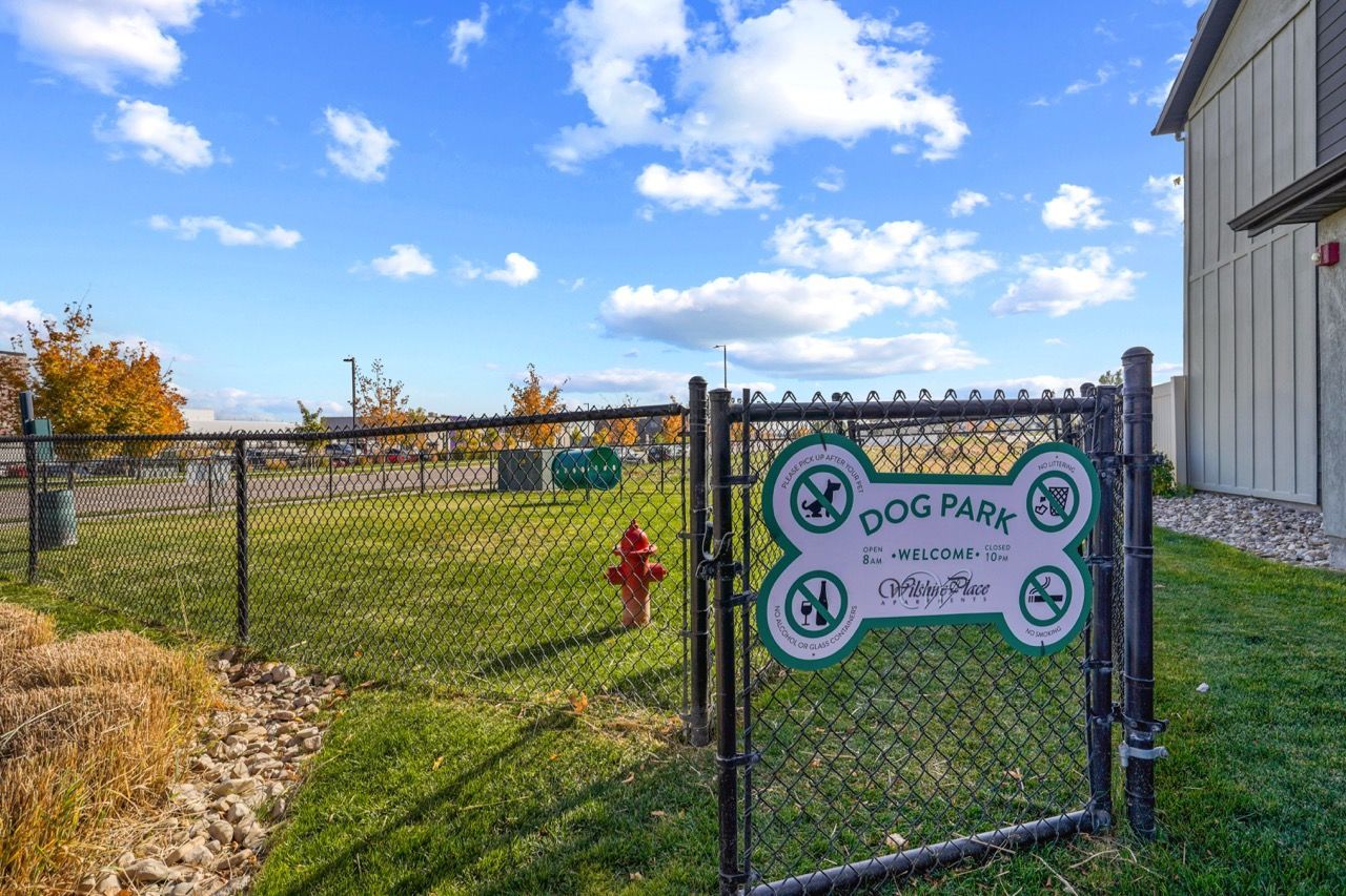 Chain-link fenced dog park gate with a large green sign beside an apartment building under a clear blue sky.