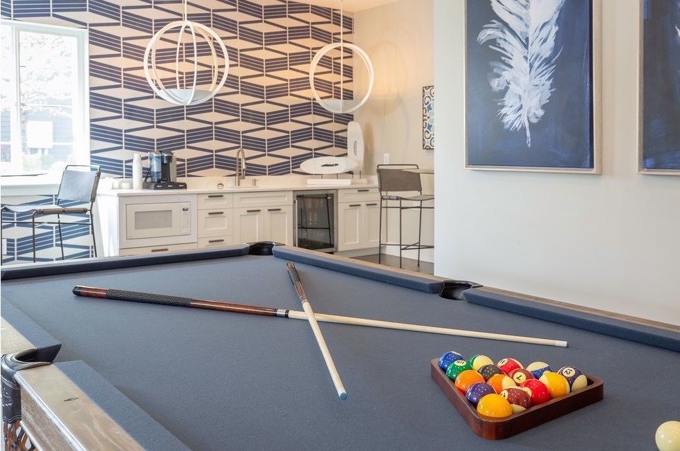 Indoor community game room with a pool table, cues, and a kitchenette in the background.