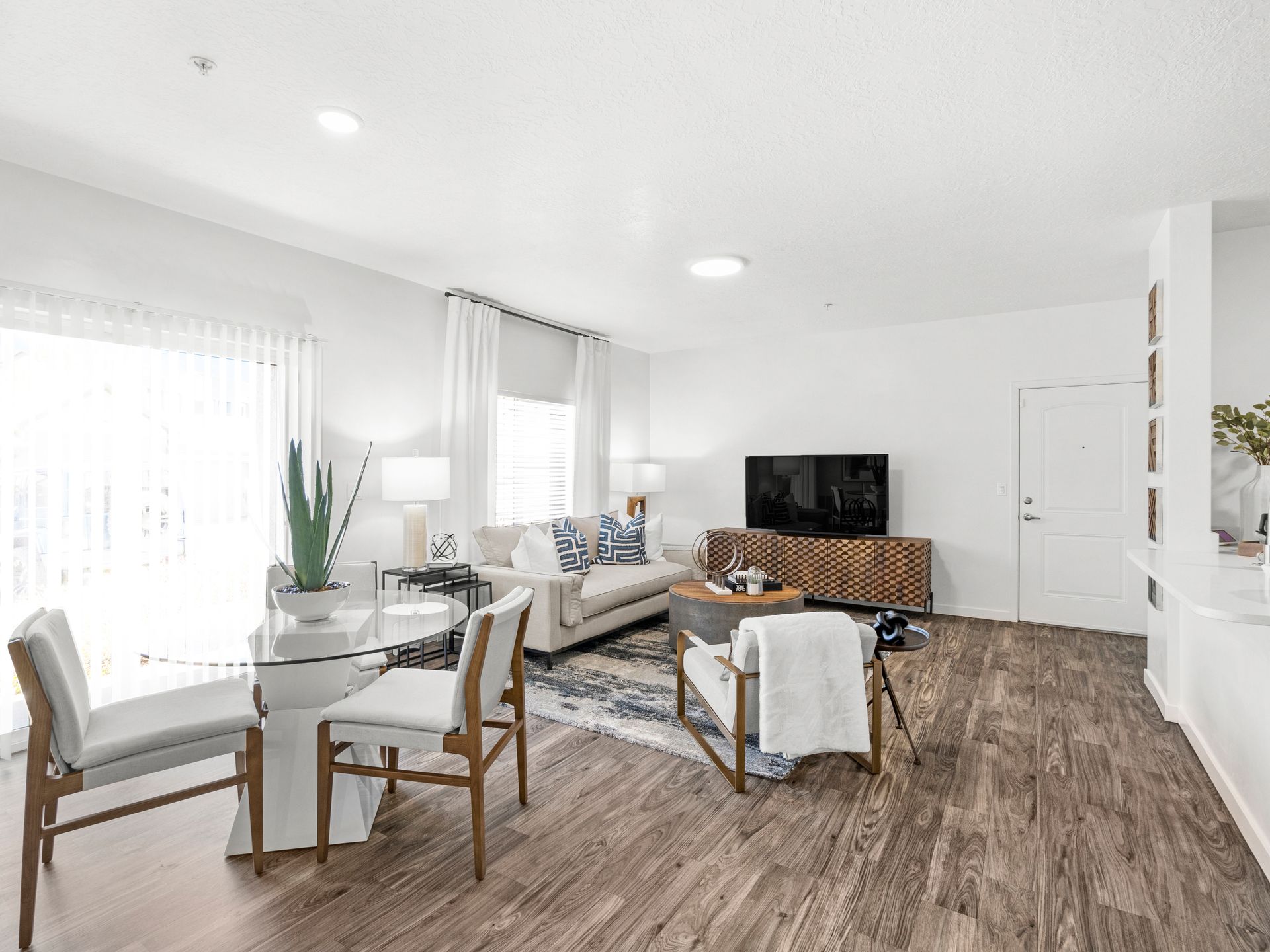 Sunlit living room with white walls, wood-look flooring, a glass dining table, and a sofa.