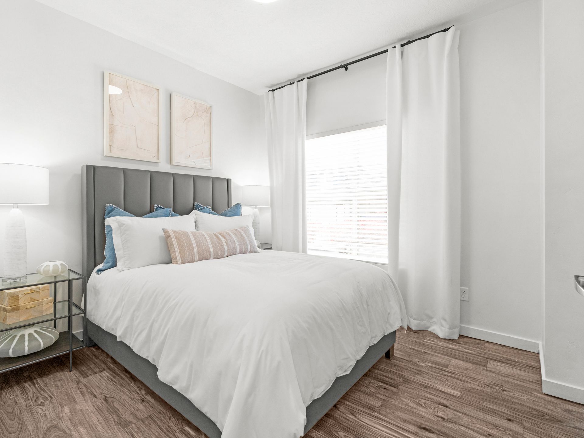 Bright bedroom in a modern apartment with a gray upholstered bed, white bedding, and white curtains.