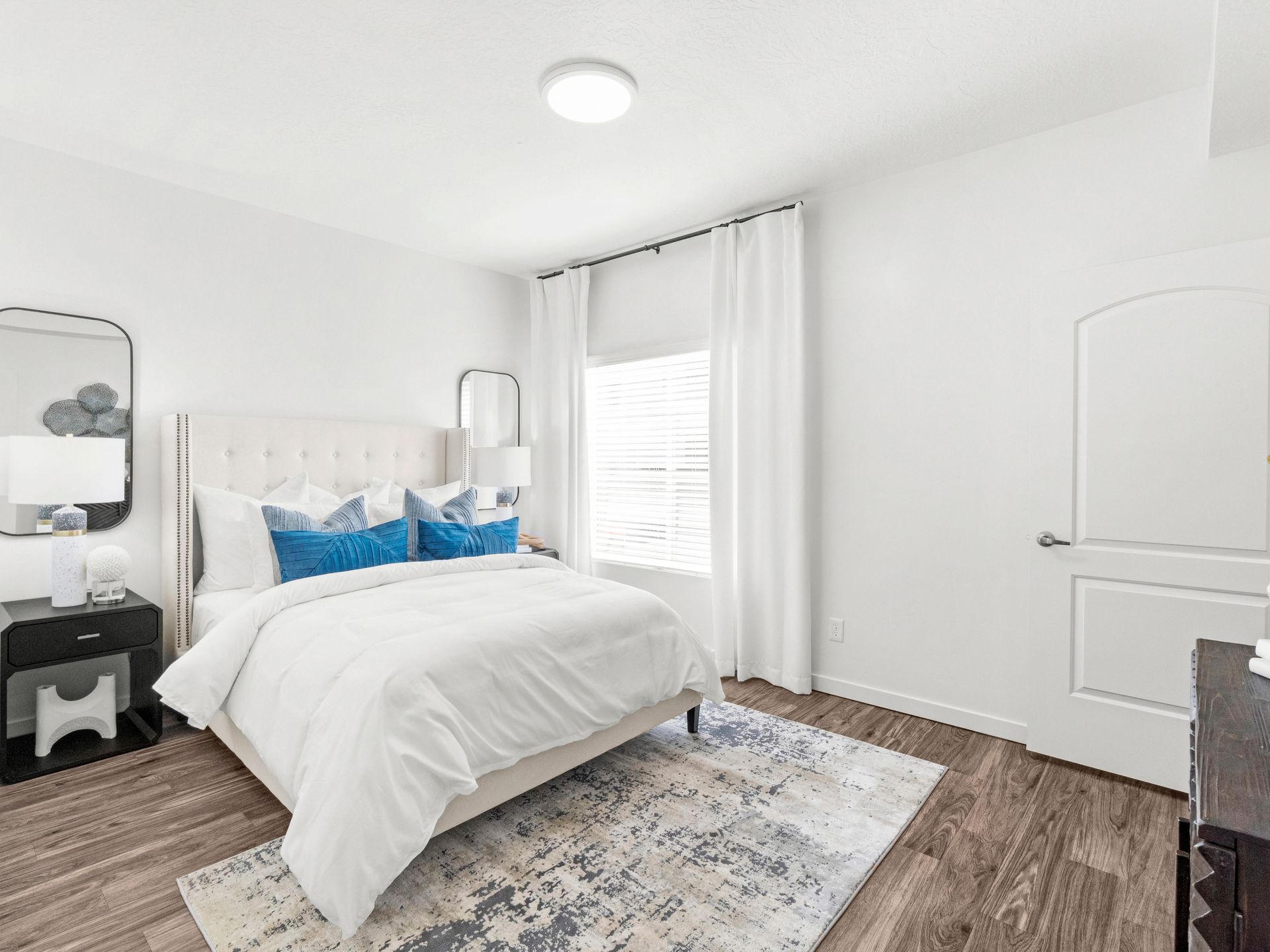 Bright white bedroom with a bed, nightstands, window with white curtains, and a patterned rug.