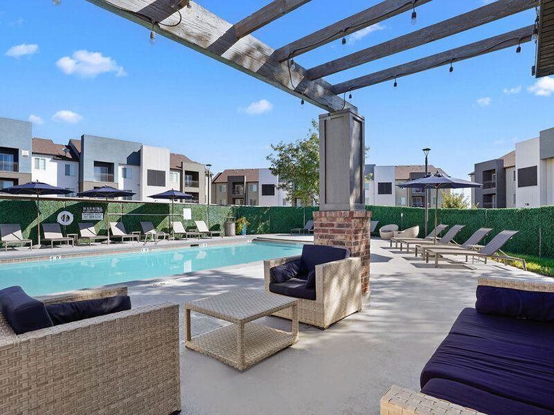 Outdoor pool area with lounge furniture under a pergola, adjacent apartment buildings.