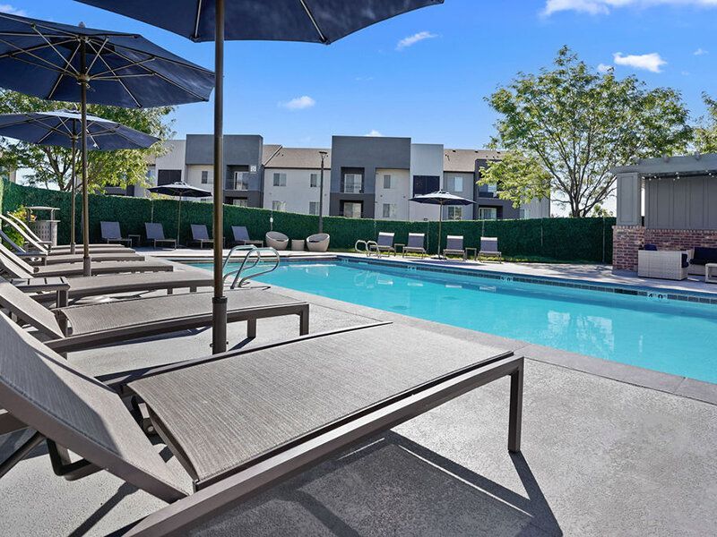 Poolside with lounge chairs and umbrellas, apartment building in background. Sunny day.