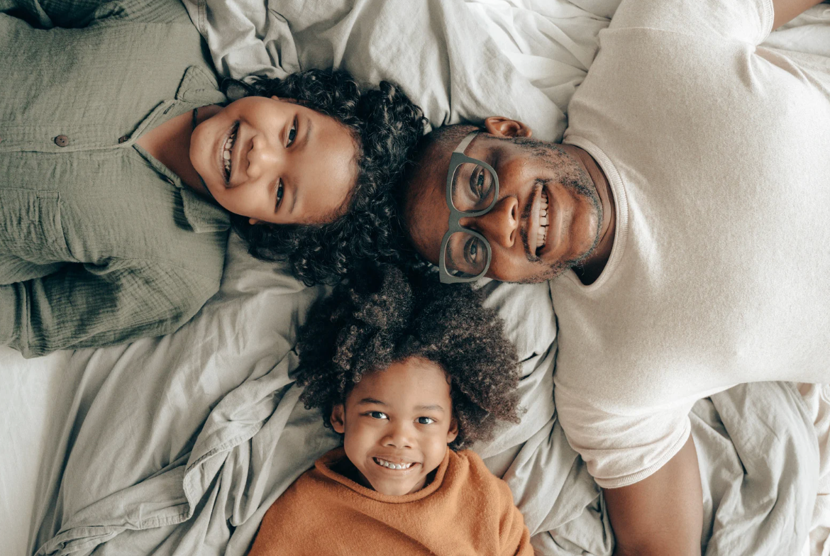 A smiling Black family lying on a bed, looking up at the camera.