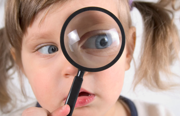 Young child looks through a magnifying glass at their eye, pigtails, blue eyes.