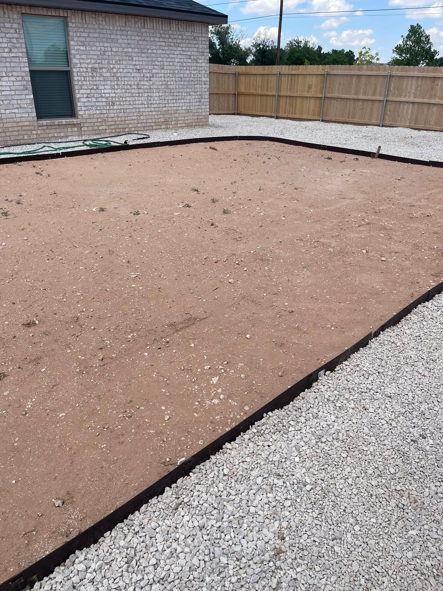 A rectangular backyard with brown dirt bordered by black edging and white gravel. A brick house and wooden fence are visible.