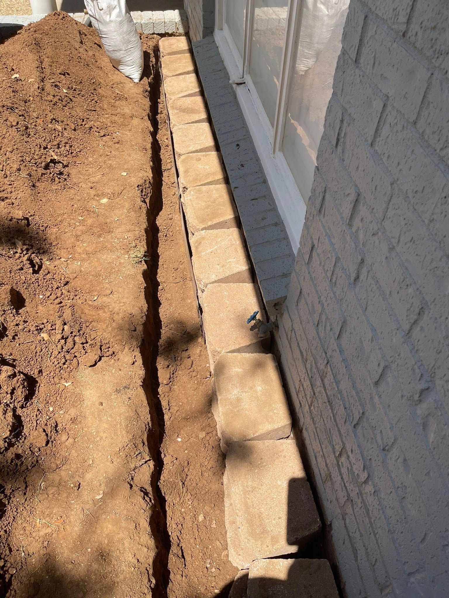 Trench dug next to a brick wall and window, with a row of paving stones along the wall's base.