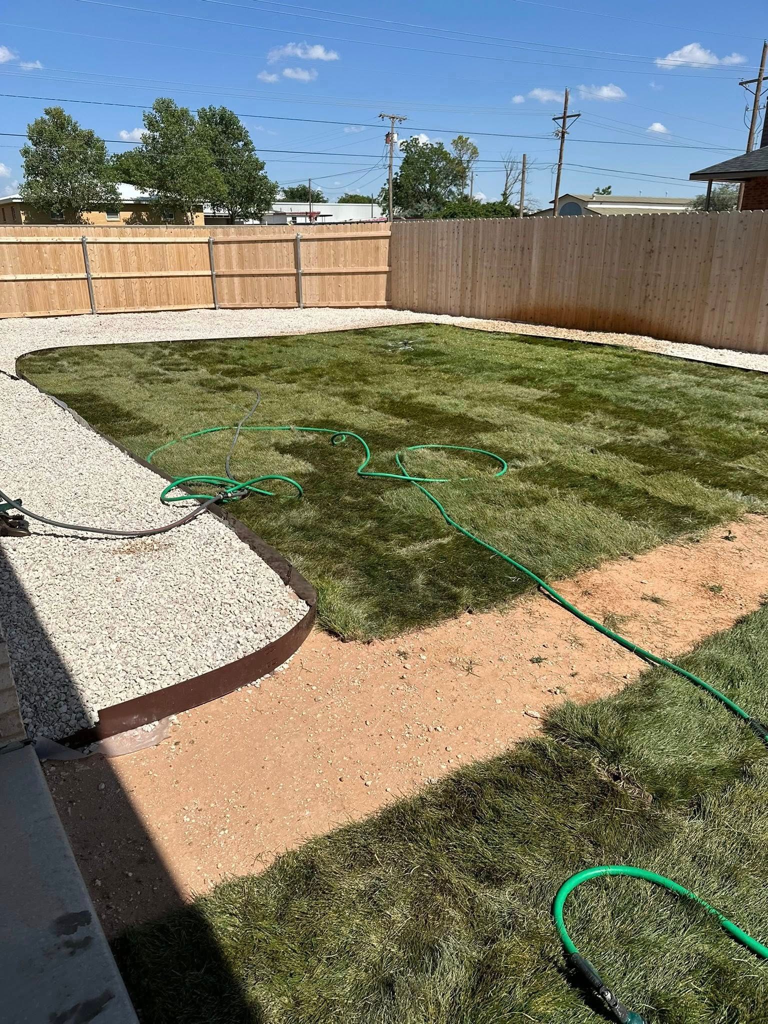 Backyard with freshly laid sod, gravel path, wooden fence, and green hose under a blue sky.